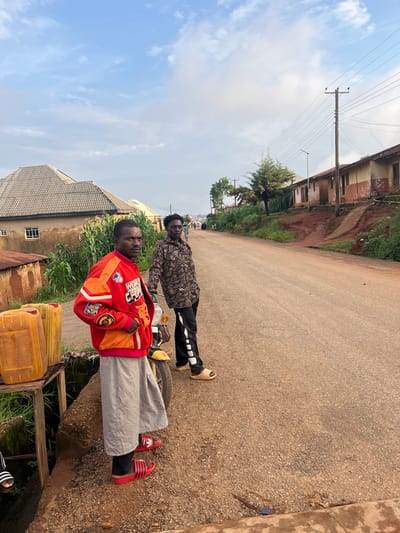 Two young men are standing on a paved road in Gembu, Nigeria. The man in the foreground is wearing a red and orange jacket with patches, grey pants, and red Adidas slides. He is looking at the camera. The man behind him is wearing a patterned shirt and black pants with white stripes, and tan sandals. He is also looking towards the camera. They appear to be near a motorcycle. In the background, there are buildings, trees, and power lines. Yellow jerrycans are visible near the front of the image. The sky is blue with some clouds, suggesting it is daytime. The overall scene depicts a common street view in a Nigerian town. The people do not display strong emotions. No visible text is present on clothing or objects.
