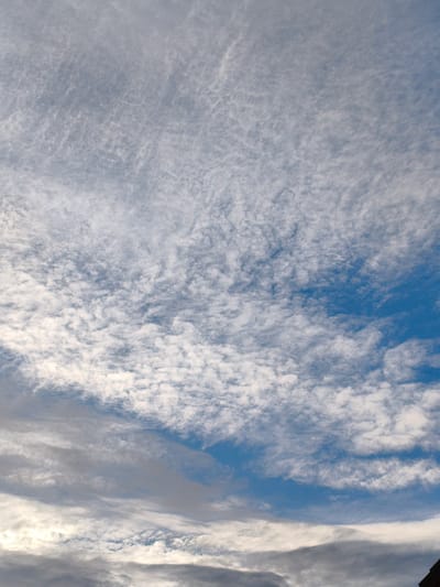 The image is a vertical, wide-angle shot of a cloudy sky above Wychavon, United Kingdom. The sky is predominantly blue, with varying densities of white and grey clouds scattered across it. The clouds range from thin, wispy cirrus formations in the upper portion of the image to thicker, more opaque stratocumulus clouds towards the bottom. The lighting suggests it is daytime, likely late morning or early afternoon, given the brightness of the clouds and the blue sky. There are no discernible people, subjects, or specific objects in the sky itself. The bottom edge of the frame shows the very top of a dark, tiled roof, possibly belonging to a building, which is the only hint of the terrestrial environment. The overall scene is natural and devoid of any human activity or artificial structures, focusing solely on the atmospheric conditions.