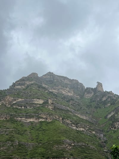 A low-angle, full shot captures a mountainous landscape under an overcast sky. The rugged, layered rock faces of the mountain are partially covered by lush green vegetation, creating a stark contrast between the barren rock and the vibrant plant life. The sky is a uniform gray, with no visible sun or distinct cloud formations, suggesting a muted or hazy atmosphere. There are no people or man-made objects visible in the frame, indicating a natural and undeveloped environment. The image appears to be a scenic view of a remote, mountainous region, likely in Assab, Russia, given the provided context.