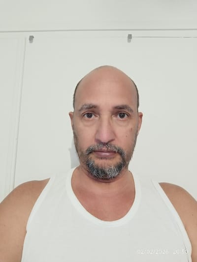 A man with a shaved head and graying beard stares directly at the camera. He wears a white tank top. The background is a plain white wall with a metallic clip visible near the top. The date "02/02/2026 20:50" is faintly visible in the lower right corner. The man's expression is neutral, and the image is well-lit with soft lighting.