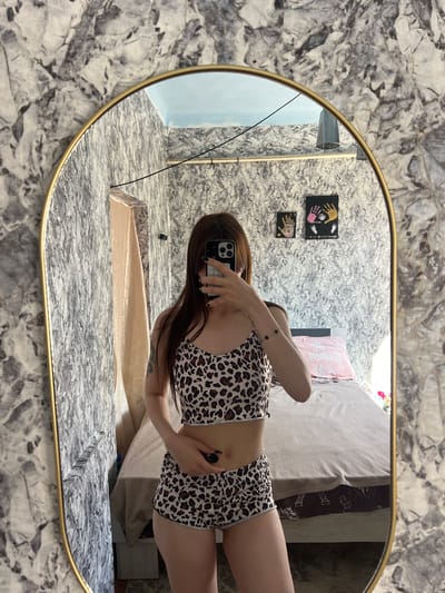 A person is taking a mirror selfie in a room with marble-patterned wallpaper. The person is wearing a leopard-print pajama set consisting of a camisole and shorts. They are holding a smartphone to take the photo. In the reflection, a bed with a pink pillow is visible, along with some framed handprints on the wall. The room appears to be a bedroom or living space. The location is stated as Blizhnii Khutor, Moldova.