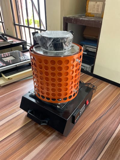 The image shows a piece of machinery with an orange, cylindrical, perforated guard surrounding a cylindrical component wrapped in plastic. The machinery is primarily black and has a red power switch. It rests on a surface with a wood grain pattern. In the background, there are glimpses of an outdoor environment and other equipment. The location is not specified, but the city is Afaka, Nigeria.