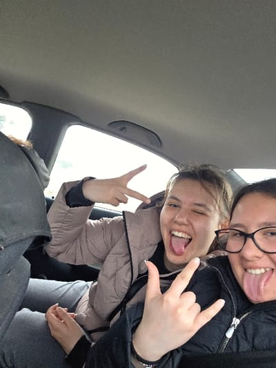 The image depicts two individuals inside a vehicle's passenger compartment, stated to be located in Muselievo, Bulgaria. Both are partially visible, seated in what appears to be the rear. The individual on the left wears a light brown puffer jacket over a dark garment and light grey pants. She presents an open mouth with an extended tongue, winks with her right eye, and displays a 'V' gesture with her raised right hand. The individual on the right, positioned in the foreground, wears black-rimmed glasses and a black zippered jacket. She also exhibits an open mouth with an extended tongue and performs a 'rock on' gesture with her raised left hand. The vehicle's interior features a light grey headliner and visible window frames, through which bright ambient light is discernible. A dark grey seat back is visible to the far left. The interaction suggests a posed photographic event.