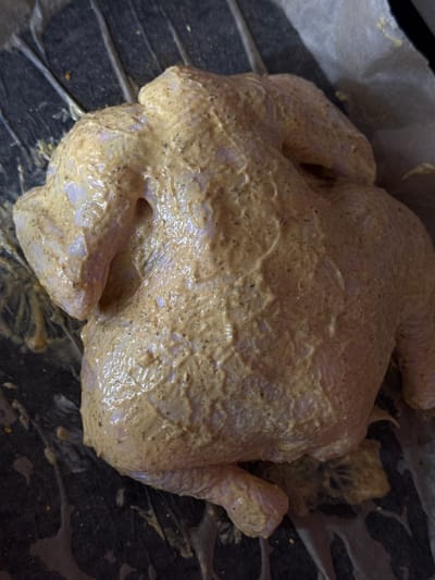 A whole chicken, coated in a yellowish marinade with visible spices, is positioned on a dark, textured surface, possibly parchment paper or a baking sheet. The chicken is fully intact and appears to be raw, with its legs and wings tucked in. The lighting is overhead, casting subtle shadows and highlighting the glossy texture of the marinade. The background is dark and out of focus, drawing attention to the prepared chicken. The image appears to be a close-up shot, focusing on the preparation of the food. There is no indication of the location being in the city of Troitske, Ukraine.