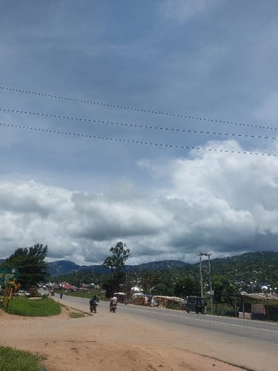 This image depicts a rural-urban road scene in Kimamba, Tanzania, under a partially cloudy sky. The perspective is from the unpaved shoulder of a two-lane asphalt road, looking forward and slightly downhill toward a distant valley framed by vegetated hills.
**Environment:**
The sky occupies approximately the upper third of the frame, displaying both blue patches and substantial white to gray cumulus clouds, indicating overcast conditions mixed with sun. Three parallel utility lines are visible horizontally across the upper sky. The foreground comprises a dirt and gravel shoulder with sparse green grass on the left. The road itself is paved with asphalt, appearing dark gray. Beyond the road, the terrain ascends into green hills densely covered with trees and scattered human settlements, including houses with red roofs. A tall, slender tree stands near the center of the mid-ground, while a larger, leafy tree is prominent on the left. Multiple utility poles with power lines are visible along the right side of the road.
**Objects and Structures:**
On the left side of the road, a green sign (partially obscured) is visible, behind which a white vehicle, possibly a truck or van, is parked. Further down the road on the left, several structures are visible amidst the trees. On the right side of the road, a series of informal market stalls or small shops with corrugated iron or tarpaulin roofs line the dirt shoulder. A dark-colored three-wheeled vehicle, resembling a rickshaw or Bajaj, is parked on this right shoulder.
**People and Actions:**
Several individuals are operating motorcycles on the road, traveling away from the viewer. One motorcycle with a rider is visible in the right lane of the mid-ground. Another motorcycle with a rider is slightly behind and to the left of the first, also moving away. A third motorcycle is discernible further down the road. No clear pedestrian figures are identifiable.