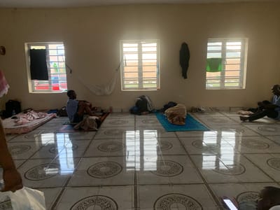 The image shows the interior of a room in Wai A, Nigeria. The room has a tiled floor with a patterned design. Three windows are visible, two on the left wall and one on the right wall. Each window has white bars for security and appears to be open. The room is sparsely furnished, with several people present.
On the left side of the room, a person is sitting on a mattress with pink bedding. To the right of this person, another individual is seated on the floor, facing away from the camera. A white hammock is suspended between the two left windows. A black bag is visible on the floor near the left wall.
In the center of the room, a person is lying down on a blue mat, covered with a beige cloth. Clothing items are scattered on the floor around them.
On the right side of the room, a person is seated on the floor, wearing a blue shirt and dark pants. This individual is facing to the left. Near the right wall, a dark garment is hanging. A green fabric is also hanging in the rightmost window.
The walls are painted a light color, and the ceiling is not fully visible. Natural light streams in through the windows, casting reflections on the tiled floor. The overall impression is of a communal living space, possibly a temporary shelter or dormitory.