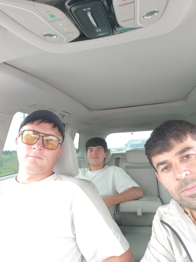 Three people are seated inside a car, taking a selfie. The person in the foreground on the left is wearing sunglasses and a black baseball cap. They are looking directly at the camera. To their right and further back is another person wearing a white t-shirt. In the background, on the right, the face of a third person is visible, looking towards the camera. The interior of the car appears to be light-colored, with a sunroof visible in the ceiling. The scene suggests they are traveling, as indicated by the reflections in the sunglasses and the car's interior. The lighting suggests it is daytime, possibly late afternoon or early evening given the warm tones in the sunglasses' reflection. No specific location cues are immediately apparent other than it being inside a vehicle.