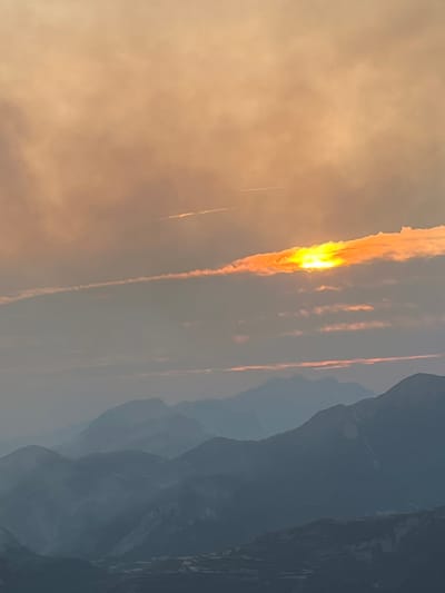 This is a photograph of a mountain range at dusk, with the sun setting behind a strip of clouds. The sky is a gradient of orange and gray, with wispy clouds and contrails visible. The mountains in the foreground are silhouetted against the sky, appearing as dark blue shapes. The overall impression is one of natural beauty and tranquility. The image is located in the vicinity of San Juan de Plan/San Chuan de Plan, Spain. There are no people, animals, or man-made structures immediately visible in the scene. The environment depicted is exclusively natural.