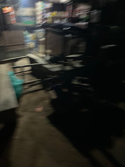The image depicts a blurry, low-light scene that appears to be an outdoor market stall. In the background, there are shelves stocked with various items, possibly packaged goods, with some colorful wrappers visible. A teal-colored plastic bag is hanging to the left, and a large dark shadow dominates the right side of the frame. The ground in the foreground is a dirt or asphalt surface, with some debris scattered on it. The overall impression is of a dimly lit, possibly nocturnal, commercial setting in what could be Safari, Nigeria.