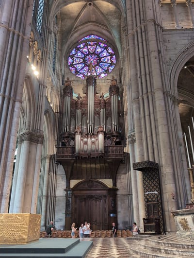 The interior of a large Gothic cathedral in Urvillers, France, is depicted. The focal point is a grand pipe organ with dark wood casing, positioned centrally against a wall beneath a large, ornate rose window filled with stained glass in vibrant blues, pinks, and purples. The architectural style is characterized by towering stone columns, ribbed vaults, and arched doorways.
In the foreground, a group of four individuals are visible standing near a golden sarcophagus or altar. One person, a man with a backpack, is looking towards the group. The group consists of three women and a man, two of the women are interacting with the man and another woman is holding a young child. Further to the right, there are several rows of empty wooden chairs.
To the right of the organ, a large, decorative banner with a fleur-de-lis pattern hangs above a wooden throne-like structure. Another person, a woman, is seated on steps near this throne. A man in a dark t-shirt is walking away from the camera on the left side of the frame, near the rows of chairs.
The floor is a checkered pattern of light and dark squares. The lighting suggests it is daytime, with natural light filtering through the windows and some artificial lighting within the cathedral. The overall scene conveys a sense of historical grandeur and religious significance.