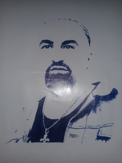 The image is a close-up, frontal, pen-and-ink sketch on a light-colored, possibly textured surface, depicting the upper body and head of a person. The sketch is done in dark blue ink and is characterized by bold, impressionistic lines.
The subject appears to be a man with a prominent beard and mustache, rendered with textured strokes to suggest hair. His eyes are wide open and looking forward, with dark, heavy brows. The nose is broadly sketched, and the mouth is a simple, dark line. He is wearing a chain around his neck, with a pendant visible. His attire includes what appears to be a collared shirt or vest, with dark lines suggesting folds and edges. The drawing style is loose and gestural, with some areas filled with denser ink to create shading, and other areas left open with sparse lines. The background is plain and light, with some scattered dots and faint lines that may be part of the drawing or imperfections on the surface. There is a noticeable sheen or reflection on the surface in the lower right quadrant of the image.
The location is specified as Gulbag, Uzbekistan. However, the image itself does not contain any visual information that would allow for identification of the specific location within Gulbag, such as landmarks, street signs, or architectural details. The drawing is a portrait and does not depict an environment or scene.