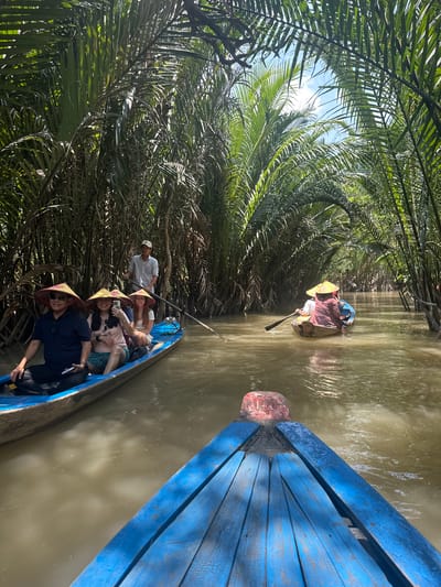 The image shows a river scene in Tân Thạch, Vietnam. A long blue boat with several passengers, some wearing conical hats, navigates the waterway. Another smaller boat with two occupants is slightly ahead. Dense vegetation lines the riverbanks. The water is murky.