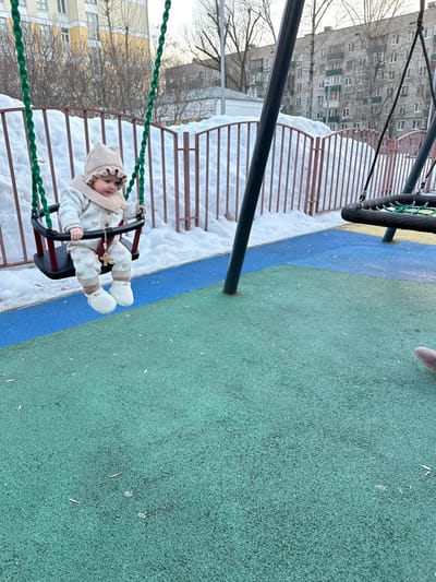 The image depicts an outdoor playground scene in Sadovaya, Russia. A young infant, dressed in a beige knitted hat, sweater, pants with heart patterns, and white booties, is seated in a red and black toddler swing with green rope chains. A small wooden cross-shaped charm is visible on the infant's attire. The swing is positioned over multi-colored rubberized playground surfacing, predominantly blue and green. Behind the swing set, a low, reddish-brown metal fence runs horizontally, separating the play area from a large accumulation of snow. In the background, leafless trees and several multi-story residential buildings with visible balconies are present under a pale sky. A partial view of an adult's foot wearing a brown shoe is visible on the green rubberized ground in the lower right corner of the frame. A second, black, circular-shaped swing is partially visible to the right of the primary swing set structure.