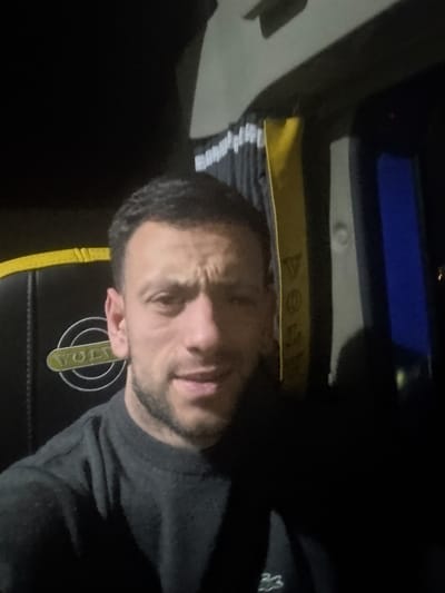 A single adult male, appearing Caucasian with short dark hair and facial stubble, is depicted in a close-up self-portrait within the interior of a vehicle cabin. He is wearing a dark, long-sleeved crew-neck shirt with a small white embroidered logo on the lower left chest area. His expression shows slightly furrowed brows and partially open lips. The background elements indicate a commercial truck interior, specifically a Volvo brand vehicle, evidenced by a prominent "VOLVO" logo on a dark panel with a yellow stripe to the subject's left. Dark curtains with white vertical stripes and yellow borders are visible to the right. A blue-tinted surface, possibly a window or display screen, is also discernible on the right side of the frame. The scene is dimly lit, suggesting night or low-light conditions. The specific city or municipality of Veria, Greece, cannot be determined from the visual information presented in the image.