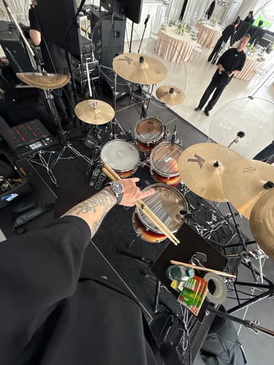 The image shows a drum set from a high-angle perspective. The drums are positioned on a black rug or platform. A person wearing a black shirt and a wristwatch is holding drumsticks over the drum set. Their arm is tattooed. There's a black table nearby with a water bottle, tape, and other items. Other people are visible in the background, standing near tables in what appears to be a venue or event space. The location is possibly Shchekutino, Russia, though this cannot be confirmed solely from the image content.