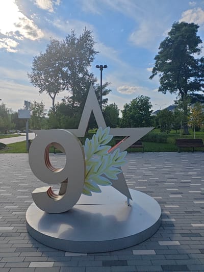 A large, metallic sculpture featuring the number "9" superimposed on a star is the central subject of this image. The sculpture appears to be made of a light-colored, possibly brushed metal, with a glossy circular base. A stylized laurel wreath, depicted with green and yellow leaves, is attached to the sculpture, adding a decorative element.
The sculpture is situated outdoors, on a paved walkway made of grey and white rectangular bricks. In the background, a park-like setting unfolds with lush green trees, well-maintained grass, and several park benches. A lamppost is visible on the left side of the frame, suggesting this is a public area. The sky above is a vibrant blue, dotted with white, fluffy clouds, and the sun is shining, casting shadows from the sculpture and trees. The lighting indicates it is daytime, likely late afternoon or early evening given the angle of the sun.
The overall atmosphere is serene and possibly commemorative, given the prominent "9" and star, which are often associated with Victory Day celebrations in Russia. There is no visible text in the image. The location context of Novyy Oskol, Russia, further strengthens the association with patriotic holidays. There are no people visible in the image.