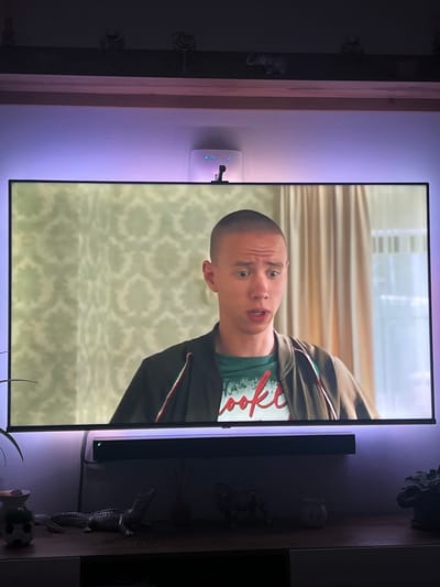 A young man with a buzz cut and a surprised expression is shown on a television screen. He is wearing a dark jacket over a green t-shirt with white text. The background on the screen appears to be a room with patterned wallpaper and curtains. The television is mounted on a wall above a dark entertainment stand. A soundbar is positioned on the stand directly below the television. The scene is illuminated by purple LED lights behind the television. On the entertainment stand, there are several decorative objects, including a ceramic alligator figurine, a small decorative pot, and a plant. The overall setting appears to be a living room. There is no information or visual evidence to suggest that this scene is located in Böckstein, Austria.