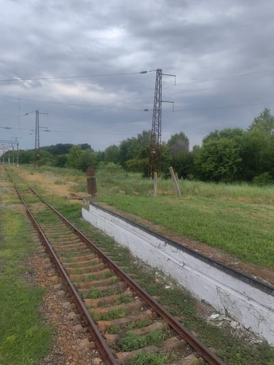 The image depicts a railway track with a platform on the right side, located in Novokubansk, Russia. The sky is overcast with heavy clouds, and lush green trees and vegetation surround the tracks. Several electricity pylons are visible in the background, with power lines stretching across the scene. The platform is made of concrete and is painted white, with some signs of wear and tear. A rusted metal signal post stands near the platform. The railway tracks themselves appear to be made of metal rails laid on concrete sleepers, with gravel ballast. The overall scene suggests a somewhat rural or semi-abandoned railway station, typical of such locations in the region. No people are visible in the image.