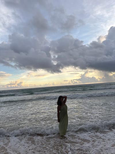 This image features a woman standing at the edge of the ocean on a beach, with waves gently washing over her feet. She is wearing a light-colored, patterned dress and has her hands behind her head, looking towards the camera with a relaxed expression. The scene is set against a dramatic sky filled with a mix of dark, heavy clouds and patches of bright, warm sunlight, suggesting a dynamic weather system. The ocean is visible to the right of the frame, with its surface reflecting the light and creating a textured pattern with the foamy waves. The overall impression is one of serene beauty, with the woman appearing comfortable and at peace amidst the natural elements. The time of day is likely late afternoon or early evening, given the quality of the light filtering through the clouds. The location is identified as Preah Sihanouk, Cambodia. There is no visible text in the image.