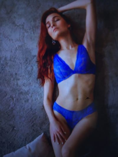 The image shows a woman with long red hair wearing blue lingerie, leaning against a textured gray wall. Her right arm is raised behind her head, and her left hand is resting on her hip. The woman is positioned in the center of the frame, with the wall occupying the background. The scene appears to be indoors, possibly a studio setting, given the plain background and soft lighting. There is no indication of the city of Dobra Krynytsia, Ukraine, or any other specific location.