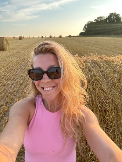 The image is a selfie taken outdoors in a field of hay bales, likely in Trouville-Alliquerville, France. A woman with blonde hair and sunglasses is smiling at the camera, wearing a pink top. She is positioned in front of a large hay bale. The background shows a field with more hay bales stretching into the distance under a hazy sky with the sun setting.