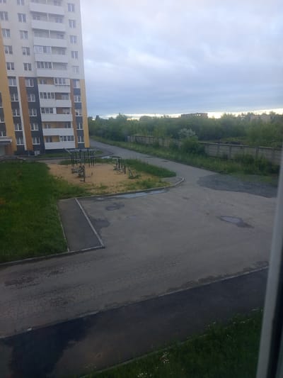 The image depicts an outdoor scene on an overcast day, likely in Kopeysk, Russia. In the foreground, a paved area with cracked asphalt shows several puddles. To the left, a grassy slope leads to a sandy playground with exercise equipment and a small jungle gym. Further back, a tall, multi-story apartment building with cream, yellow, and grey facades stands prominently. Beyond the apartment building, a row of trees forms a green barrier, behind which a concrete fence is visible. In the distance, several dilapidated brick buildings, possibly industrial or abandoned structures, are partially obscured by vegetation. A dark-colored car is parked on the left side of the image, near the apartment building's entrance. The sky is cloudy, with soft light filtering through.
