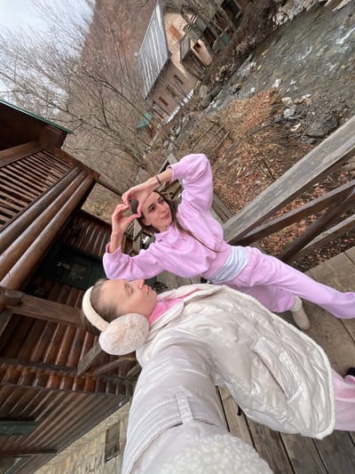 The image captures a high-angle, selfie-style perspective taken on a wooden structure, likely a porch or bridge, located in Crkvine, Montenegro. In the foreground, partially visible, is an individual wearing a light-colored puffer jacket and fluffy cream-colored earmuffs, appearing to hold the camera. Their eyes are closed or looking downwards. Behind this individual, a second female with long dark hair stands, wearing a pink hooded tracksuit over a white top. She is engaged in the action of forming a heart shape with her hands, extending her arms outwards, and looking towards the right of the frame. The immediate environment includes a rough-hewn wooden wall to the left, and visible wooden planks and railings of the structure. The background reveals a narrow, rocky river or stream flowing below. Across the river, multiple rustic buildings with varying rooflines and wooden balconies are visible amidst sparse, bare deciduous trees and dry brown foliage, characteristic of a colder season. Beyond the river and buildings, the landscape consists of steep, deforested mountainous slopes under a uniform, overcast sky.