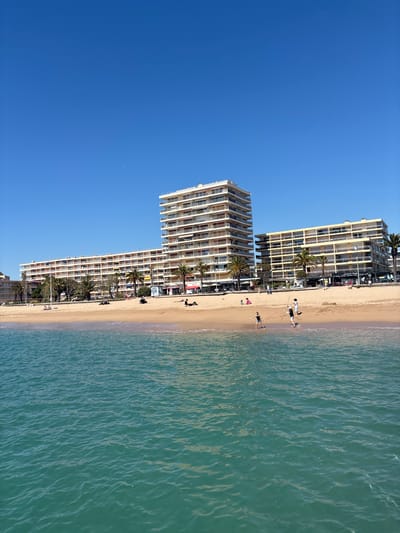 The image depicts a beach scene in Fréjus, France. Tall, multi-story apartment buildings line the shoreline. The sand is light beige. People are visible on the beach, some walking near the water's edge and others sunbathing or relaxing. The sea has a turquoise-green hue with subtle ripples. The sky is clear and blue.