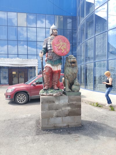A full-shot color photograph captures a statue of a warrior and a lion in front of a modern building with a glass facade. The warrior statue stands tall, clad in chain mail, a helmet, and red boots, holding a shield with a lion emblem. Beside him, a lion statue is posed with its paw on a ball. A red SUV is parked to the left of the statues. A blonde woman in a black tank top and jeans is standing on the sidewalk to the right, looking at her phone. The background features the reflective glass exterior of a building, showing clouds and the surrounding environment. The scene is set in Chesnokovka, Russia, likely in front of a commercial or public building.