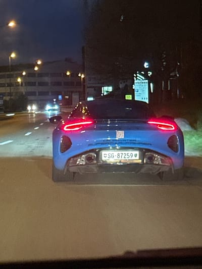 The image displays a nighttime street scene in Volketswil, Switzerland. A bright blue sports car is centrally positioned, viewed from its rear, occupying the right lane of a wet, multi-lane asphalt road. Its red LED taillights are illuminated. A white Swiss license plate, bearing "SG-87259", is mounted centrally on the rear bumper. Below the license plate, two circular exhaust pipes are visible, and discernible text on the bumper includes "HP. PRIVATE" and "SUPERCAR-PROPER". The road reflects numerous yellow-orange streetlights overhead and the headlights of several distant vehicles in the opposing or left lanes. To the left, a multi-story commercial or industrial building is brightly illuminated by multiple streetlights. To the right, a white sign with blue text, partially reading "wipfass" and displaying a blue 'P' (parking) symbol, is visible amidst dark vegetation. No individuals are present in the frame.