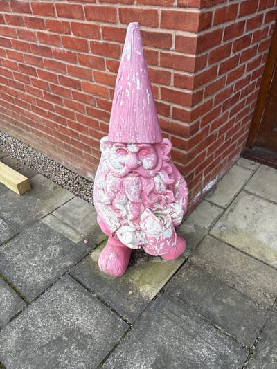 An outdoor ground-level shot shows a pink and white painted statue of an ice cream cone resting against the brick wall of a building in the city of Shrewsbury, United Kingdom. The statue is positioned horizontally in the frame. The cone section points towards the left and the multiple scoops of ice cream extend to the right. The paint is faded and chipped, revealing the base material in places. The wall is constructed of red bricks with light-colored mortar and forms a step or platform alongside the building. Adjacent to the wall is a paved area composed of gray concrete slabs. Between the brick wall and concrete slabs is a narrow strip of gravel, and a piece of wood lies nearby. The image is taken in daylight, and the environment appears to be an exterior location.
