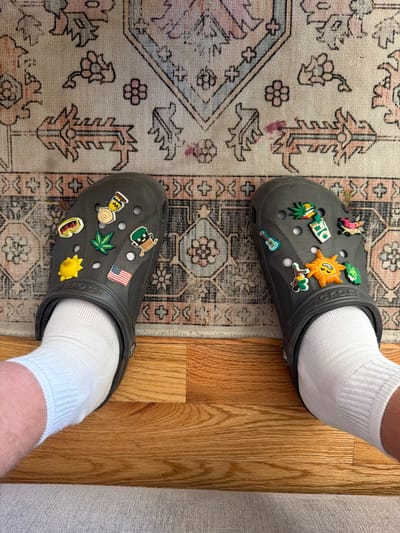 The image depicts a person wearing gray Crocs with white socks standing on a patterned rug and wooden flooring, in an indoor setting. The Crocs are adorned with various decorative charms. One Croc has a charm resembling a sun and the other has a charm resembling the American flag. The flooring appears to be hardwood. The location in Morton Grove, United States of America is not determinable from the image alone.