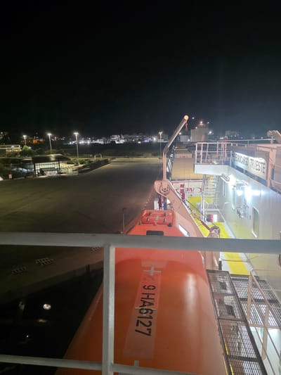 The image is a night time photograph taken from the deck of a ship docked in Brindisi, Italy. The foreground shows a metal railing and the orange hull of a life raft with the registration number "9 HA6127" printed on its side. Behind the life raft, a section of the ship's superstructure is visible, including a crane and the name "EUROCARGO TRIESTE" emblazoned on the side. In the middle ground, a paved area, likely a dock or ferry terminal, is illuminated by bright streetlights. Several buildings and structures are visible in the background, as well as distant city lights, indicating an urban or port area. The overall impression is of a commercial port at night.