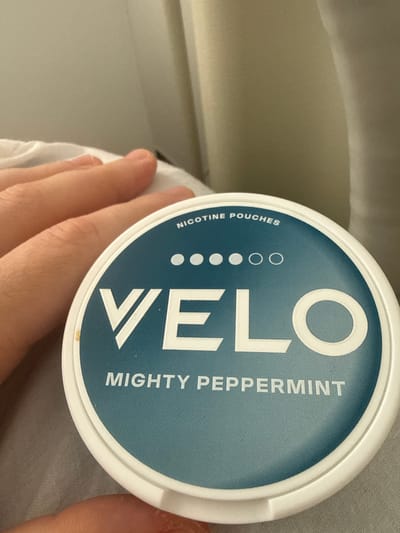 A close-up shot depicts a white, circular container with a blue label for "Velo Mighty Peppermint" nicotine pouches. The container is held in a person's hand. The background is blurred, showing a light-colored surface and a glimpse of a white material, possibly fabric. There is no indication of the location in Vynnyky, Ukraine, or any other city.