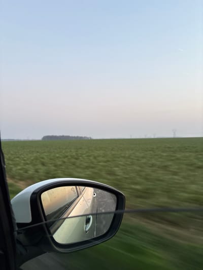The image displays a view from inside a moving vehicle, looking out the passenger side window and incorporating a portion of the vehicle's white exterior mirror. The mirror, positioned in the lower left quadrant, reflects the car's bodywork, including a visible door handle, and the blurred landscape behind. The dominant feature of the midground is a vast expanse of green agricultural field, rendered with motion blur indicative of forward movement. On the distant horizon, a dense line of dark green trees is visible on the left, while a series of tall, grey electrical pylons stretches across the right. The sky above presents a pale gradient, transitioning from a light blue at the top to a faint pinkish-grey near the horizon line. A subtle white contrail is discernible in the upper right corner of the sky. The scene depicts a rural environment near Bû, France.