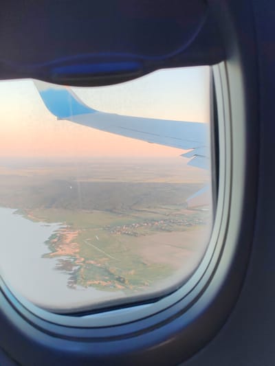 The image is taken from an airplane window, looking out at a landscape during what appears to be sunset or sunrise. The wing of the airplane is visible in the foreground, with its white surface and a blue tip. Below the wing, a body of water borders a landmass covered in green vegetation and some scattered buildings. The sky is a soft gradient of orange, pink, and yellow. The overall impression is one of peaceful travel over a scenic rural area. The description of Khrabrovo, Russia, is not supported by the visual information in the image, as there are no specific landmarks or identifiers that confirm this location.