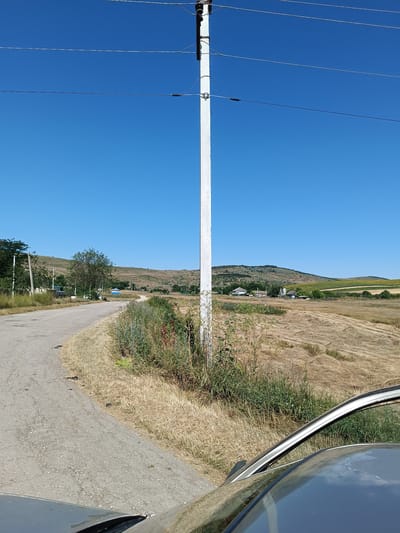 A grey car's hood and windshield frame the foreground of an image taken in Brejeni, Moldova. The car faces a rural scene dominated by a bright blue sky. A tall, white utility pole with power lines stretches vertically near the center of the frame. To the left, a paved road curves away into the distance, bordered by dry, yellowed grass and patches of green vegetation. Beyond the road, a landscape of rolling hills unfolds. The hills are covered in sparse green trees and dry grass. In the middle ground, a few houses with tiled roofs are visible, nestled among trees. The fields in the middle and background appear to have been recently harvested, with swathes of dried hay or stubble. The overall impression is one of a sunny, dry day in a rural agricultural area.
