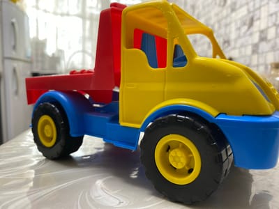 A brightly colored plastic toy truck, predominantly yellow, blue, and red, sits on a table with a patterned surface. The truck is a model of a towing vehicle, featuring a yellow cab with blue interior seats and a red rear section. It has large black tires with yellow rims. In the background, a white refrigerator is visible, along with a window with white curtains, suggesting an indoor setting. The overall scene is likely in a residential area within Бескөл ауылдық округі, Kazakhstan, given the context. There are no people or animals present in the image.