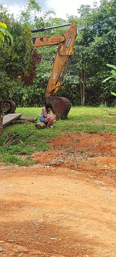A woman and a child are sitting on the grass next to a large yellow excavator. The woman is wearing a light blue tank top and denim shorts, and the child is wearing a pink shirt and light-colored pants. The excavator has a large metal bucket and a long arm extended. The scene is outdoors, with lush green trees and foliage in the background, and a dirt path in the foreground. The location is stated to be in the city of Xayden, Laos.