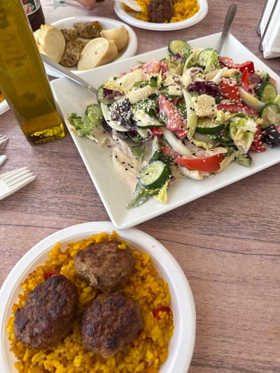 The image shows a close-up of a table setting, likely in Foia L'om, Spain, featuring a meal. The primary subjects are two plates of food and a bottle of olive oil.

The plate in the foreground contains yellow rice topped with three oval-shaped meatballs. The rice appears to be seasoned with red flecks, possibly bell peppers.

To the upper right, a white rectangular plate holds a colorful salad composed of lettuce, sliced cucumbers, tomatoes, red onions, and possibly cheese cubes, all coated in a creamy dressing and sprinkled with sesame seeds. A fork is resting in the salad.

To the left, a tall, clear glass bottle of olive oil is positioned on the wooden table. Next to the oil bottle, a white plate holds several pieces of bread, some appearing to be sliced baguettes and others darker, possibly toasted or whole-wheat bread.

The background is softly blurred, suggesting an outdoor or casual dining environment. The wooden texture of the table is visible throughout the image. The overall scene depicts a prepared meal ready to be eaten.