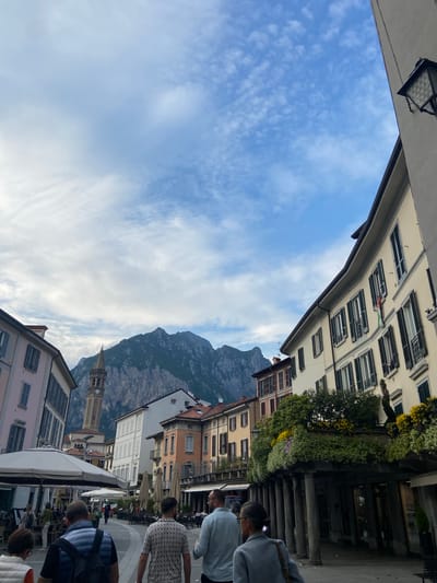 Captured at a rotated angle, the image presents a street scene in Lecco, Italy. The sky, filled with white-streaked clouds against a blue backdrop, is framed by buildings on both sides. The architecture ranges from buildings with shutters and flower boxes to a church tower. In the background, a mountain rises majestically. Pedestrians stroll through the street, suggesting a lively atmosphere. Street-side cafes and shops line the road, and flags of Italy can be spotted. The focus seems to be on the blend of urban life against the natural beauty of Lecco.