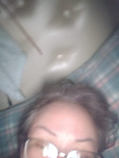 The image is a blurry, poorly lit close-up shot of a person's face and upper body, taken from a low angle. The subject appears to be wearing glasses. The background is indistinct, with a plaid-like pattern visible in some areas. There is no discernible information about the location in At-Bashy, Kyrgyzstan, or any specific actions or interactions taking place.