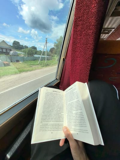 The image shows a person's hand holding an open book, with the text visible on the pages. The setting appears to be inside a vehicle, likely a bus, as suggested by the red upholstery and the window view. Outside the window, a rural landscape is visible with houses, trees, a road, and a cloudy blue sky, indicating daytime. The book is in Russian, and the visible text includes page numbers "34" and "35" along with Russian words and phrases. The overall mood is one of quiet contemplation and relaxation, with the person engrossed in reading while traveling through a scenic, possibly Russian, countryside.