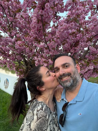 A couple is posing for a photo under a blooming cherry tree. The man, with a beard and blue shirt, is smiling. Sunglasses are tucked into his shirt. The woman, with her hair in a ponytail and wearing a snakeskin-patterned top, is kissing him. The location appears to be in Grevenmacher, Luxembourg, indicated by a sign with pictures visible in the background. The setting suggests a park or public space.