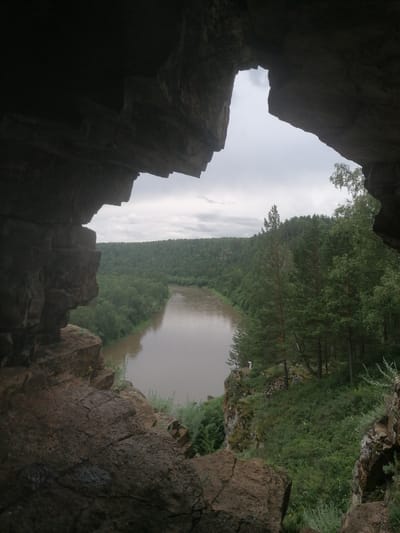 This is a medium shot, taken from within a rock overhang, looking out at a wide, calm river that curves through a dense, green forest. The sky is overcast and grayish. The foreground is dominated by rough, textured rock formations that frame the view of the river and forest. The river is a murky brown color, and its banks are lined with lush trees, primarily evergreens, with some deciduous trees interspersed. The forest extends as far as the eye can see, covering rolling hills. There are two small figures visible on the riverbank to the right of the image, standing near the edge of the water and looking towards the forest. The scene depicts a natural landscape, likely a scenic overlook or a viewpoint near a river in a forested area.