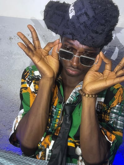 A young man with dark skin wears a distressed black cap, silver-framed sunglasses, and a patterned shirt with green, black, and yellow hues. He's also wearing a wooden beaded bracelet. He holds his hands up near his face in a stylized pose, fingers spread. The background features a white wall and a glimpse of light blue.