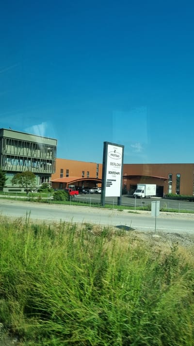 The image captures a commercial or industrial area on a clear, sunny day, with a bright blue sky dominating the upper portion. In the foreground, lush green grass and foliage are visible, suggesting a roadside setting.
The middle ground features a road and a few parked vehicles. To the left, a modern, multi-story building with a gray facade and extensive glass windows stands prominently. Adjacent to it, to the right, is a long, single-story building with an orange-brown facade and a curved, covered entrance area. A white pylon sign is positioned in front of the orange-brown building, displaying several brand names. These include "PROTEAS" at the top, followed by "BERLONI" and "BONTEMPI" below. A white delivery van with a logo that appears to be "PROTEAS" is parked near the building entrance. A red pickup truck is also visible further back, parked near the covered entrance.
The scene appears to be located within the Municipal Unit of Larissa, Greece, given the context. The overall impression is one of a business park or industrial estate situated along a roadway, bordered by natural greenery.