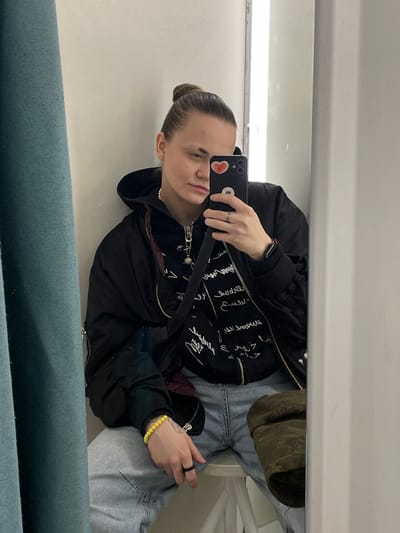 A person with light skin and hair pulled back in a bun takes a selfie in a mirror. They are wearing a black jacket over a shirt with white writing and light blue jeans. Accessories include a yellow bracelet, a black ring, a pearl necklace, and a crossbody bag with a black strap. The phone case has a heart sticker. The mirror reflects a white wall and a blue-green surface in the foreground.