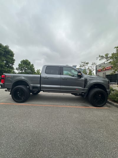 A gray Ford F-series Super Duty pickup truck is parked in a lot. The truck is a crew cab model with a long bed and has black rims and off-road tires. The truck is positioned at an angle, with its left side facing the viewer. The background shows a building with the words "Burnaby Harley-Davidson" and "8859" visible on its facade. There are trees and an overcast sky present. The location is identified as Township of Langley, Canada.