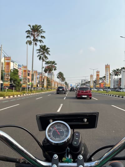 The image is a first-person perspective shot from a motorcycle in motion on a road in Pakulonan Barat, Indonesia. The foreground shows the motorcycle's handlebars, a speedometer with a red needle pointing to approximately 50 km/h and an odometer reading 0915 km, and a neutral (N) indicator light. The road ahead is paved with visible lane markings and is lined with palm trees and vertical banners advertising businesses, some of which read "FINORE" and "OPEN SHOW UNIT". Several cars are visible on the road, including a red hatchback and a black SUV. The sky is clear and bright, suggesting daytime. Buildings and other vehicles are visible in the distance, indicating an urban or suburban environment.